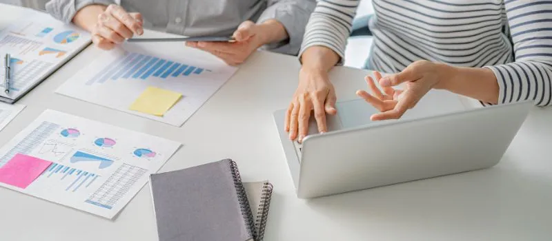 Two people reviewing printed bar and pie charts next to an open laptop during a data review meeting